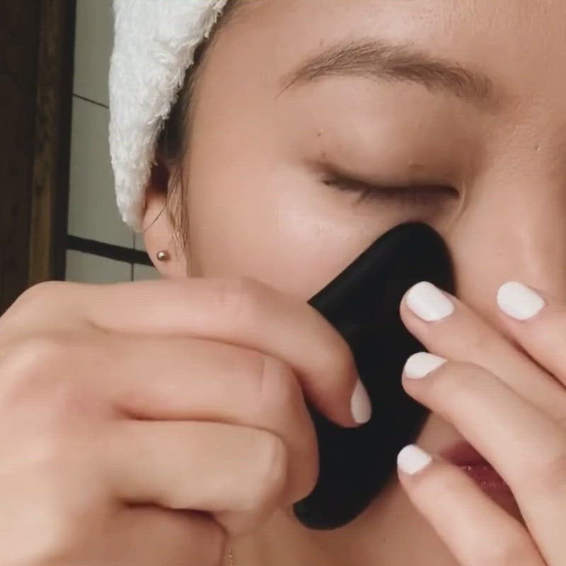 Load video: woman using award winning black obsidian gua sha lifting tool along cheeks for de-puffing